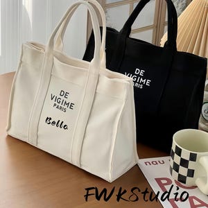 Könnte beinhalten: Zwei Canvas-Tragetaschen, eine cremefarbene und eine schwarze, mit weißen Buchstaben. Auf der cremefarbenen Tasche steht "DE VIGIME PARIS Bella". Auf der schwarzen Tasche steht "DE VIGIME PARIS". Beide Taschen haben stabile Griffe.