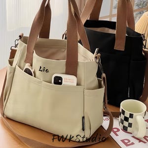 Puede incluir: Dos bolsos de mano de lona, uno crema y otro negro, con asas y correas marrones. El bolso crema tiene múltiples bolsillos, que contienen gafas de sol y un teléfono. El texto "Lia" es visible en el bolso. Una taza con un patrón de cuadros blancos y negros está cerca.