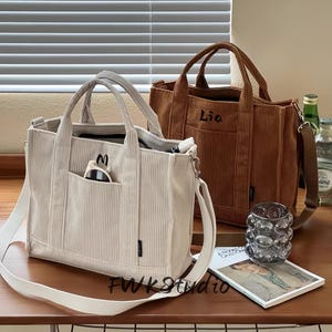 Könnte beinhalten: Zwei Cord-Tragetaschen in Creme und Braun stehen auf einer Holzoberfläche. Die cremefarbene Tasche hat eine Vordertasche und einen weißen Schultergurt. Die braune Tasche hat einen oberen Griff und den Namen "Lio" aufgestickt. Eine Sonnenbrille ist in der Tasche der cremefarbenen Tasche zu sehen.