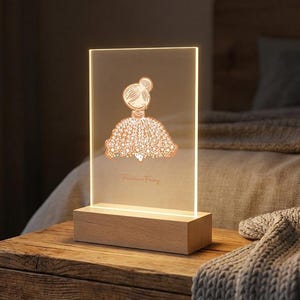 Puede incluir: Lámpara LED rectangular con base de madera. La lámpara presenta un dibujo de una mujer con un vestido. El texto "Fashion Fairy" está en la parte inferior de la lámpara. La lámpara está sobre una mesita de noche de madera.