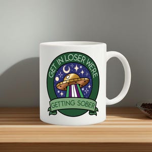 May include: White ceramic mug with a green and blue graphic of a UFO, stars, and the text "GET IN LOSER WERE GETTING SOBER". The mug has a curved handle and is sitting on a wooden surface.