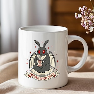Puede incluir: Taza de cerámica blanca con un diseño de criatura tipo polilla de dibujos animados con ojos rojos, sosteniendo una taza de café. La taza tiene el texto "TRUST THE PROCESS" y "EMOTIONAL SUPPORT CRYPTID". El diseño es en tonos grises, negros y rojos.