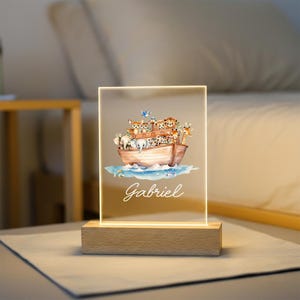 May include: A decorative night light featuring a watercolor illustration of Noah's Ark with various animals. The light is mounted on a wooden base and has the name "Gabriel" written in cursive. The light is rectangular and emits a soft glow.