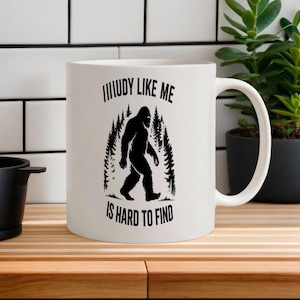 Puede incluir: Taza de cerámica blanca con una silueta negra de Bigfoot caminando por un bosque. La taza tiene el texto "MUUDY LIKE ME IS HARD TO FIND" en negro. La taza está sobre una superficie de madera.