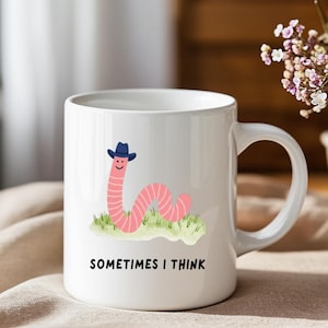 May include: White ceramic mug featuring a cartoon worm wearing a blue cowboy hat, with the text "SOMETIMES I THINK". The worm is pink and has a smiling face. The mug has a curved handle and is set against a neutral background.