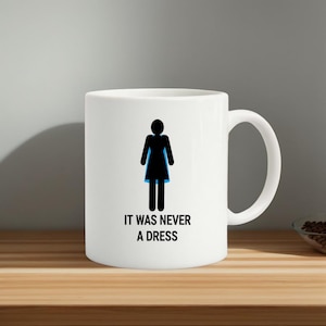 Könnte beinhalten: Weiße Keramiktasse mit einer schwarz-blauen Grafik einer Person. Der Text "IT WAS NEVER A DRESS" ist unter der Grafik aufgedruckt. Die Tasse hat einen großen, gebogenen Henkel.