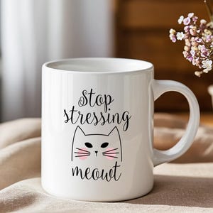 May include: White ceramic mug with the text "Stop stressing meowt" and a cat face illustration. The cat has pink cheeks and whiskers. The mug has a curved handle and is sitting on a beige surface.