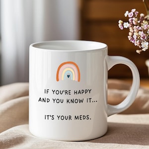 Puede incluir: Taza de cerámica blanca con un diseño de arcoíris y el texto "IF YOU'RE HAPPY AND YOU KNOW IT... IT'S YOUR MEDS." El arcoíris presenta arcos naranjas, amarillos, azules y rosas con un pequeño corazón azul. La taza tiene un asa curva.
