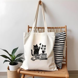 May include: Cream-colored tote bag with a black cat illustration. The cat sits atop a stack of books, with a plant in a teacup and a four-leaf clover. The bag has long handles and is hanging on a wooden hook.