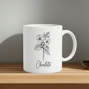 Puede incluir: Taza de cerámica blanca con una ilustración floral y el nombre "Charlotte" en escritura cursiva. La taza tiene un acabado moteado y un asa curvada.