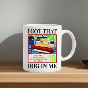 Puede incluir: Taza de cerámica blanca con asa, con un gráfico colorido de un perrito caliente, condimentos y un vaso de Pepsi. El texto en la taza dice "I GOT THAT DOG IN ME" e incluye el precio.