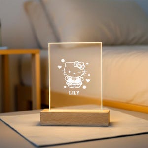 May include: A rectangular LED night light with a wooden base. The light features a white etched design of a cartoon cat with a bow, hearts, and the name "LILY". The light is on, illuminating the design.