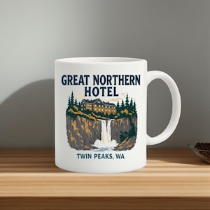 Może przedstawiać: Biały ceramiczny kubek z napisem "GREAT NORTHERN HOTEL" i ilustracją hotelu na klifie z wodospadem. Tekst "TWIN PEAKS, WA" znajduje się poniżej. Kubek ma zakrzywiony uchwyt.