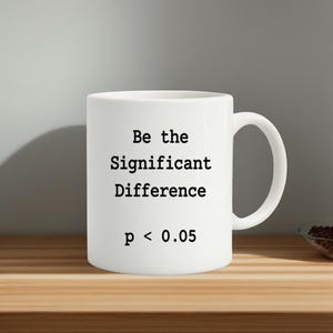 Puede incluir: Taza de cerámica blanca con el texto "Be the Significant Difference p < 0.05" en negro. La taza tiene un asa curva y está sobre una superficie de madera.