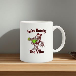 May include: White ceramic mug featuring a frog illustration with a martini and disco ball. The mug has the text "You're Ruining The Vibe" in maroon lettering.