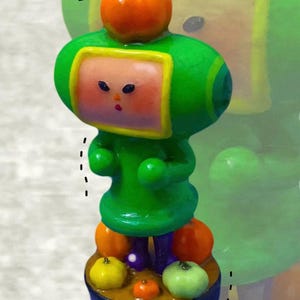 Puede incluir: Una pequeña figura que representa un personaje verde con cabeza cuadrada, vestido con un traje verde y coronado por una calabaza naranja. El personaje se apoya sobre una base circular decorada con varias calabazas pequeñas de color naranja, amarillo y verde claro.
