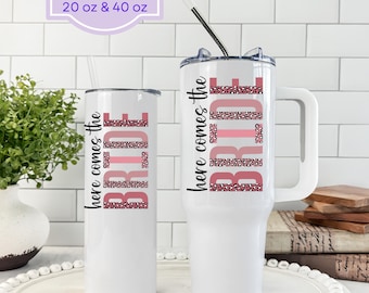 May include: White tumblers with the text "here comes the BRIDE" in pink and leopard print. One tumbler is tall and slender, the other is a large mug with a handle. The text "Tumblers available in 20 oz & 40 oz" is in the top left corner.