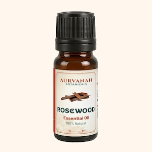 Peut inclure: Un petit flacon en verre ambré avec un bouchon noir strié contenant de l'huile essentielle de bois de rose Aurvanah Botanicals. L'étiquette blanche affiche le nom de la marque, un graphique de copeaux de bois, Rosewood Essential Oil et 100% Natural.