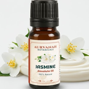 May include: A brown glass bottle with a black cap, labelled "JASMINE Absolute Oil 100% Natural" by AURVANAH BOTANICALS. The label features a jasmine flower illustration. White jasmine flowers and a creamy white substance are in the background.