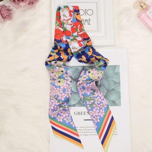 May include: A long, narrow silk scarf featuring a vibrant floral pattern with red, orange, and white blossoms on a dark blue background at the top, transitioning to a light blue section with pink flowers and a multi-colored striped border at the ends.