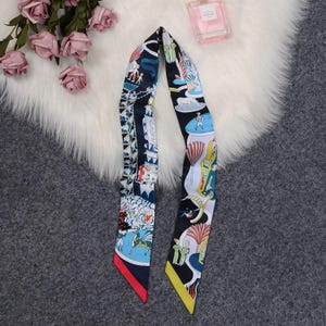 May include: A long, narrow silk scarf featuring a dark blue and white background with a multi-colored graphic print of various figures, architectural elements, and stylized plants. The scarf has pointed ends, one finished with a red edge and one with a yellow edge.