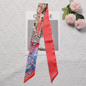 Puede incluir: Un pañuelo de seda largo y estrecho con un diseño de doble cara. Un lado muestra un leopardo, flores tropicales y pájaros en colores vibrantes. El otro lado es una tela roja brillante con un patrón de pequeños corazones azules repetidos.