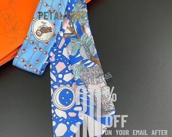 May include: A long, narrow silk scarf featuring a light blue section with a circular owl illustration and a darker blue section with a geometric pattern, a moon with stars, and botanical motifs. The scarf has a black care tag attached to the side.