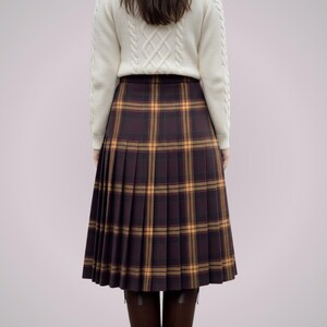 May include: A pleated, knee-length skirt in a brown and yellow plaid pattern. The skirt is paired with a cream-colored cable-knit sweater. The overall style is classic and preppy.