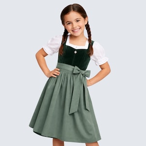 May include: A young girl in a traditional green dirndl dress. The dress has a dark green bodice, a light green skirt, and a white blouse with puffed sleeves. A green bow is tied at the waist. The girl is smiling.