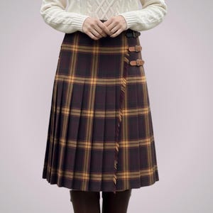 May include: A pleated, knee-length skirt in a brown and yellow plaid pattern. The skirt features a wrap-around design with leather buckle closures. The garment is worn with a white cable-knit sweater.