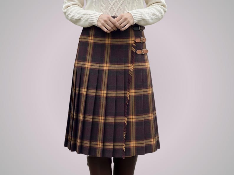 May include: A pleated, knee-length skirt in a brown and yellow plaid pattern. The skirt features a wrap-around design with leather buckle closures. The garment is worn with a white cable-knit sweater.