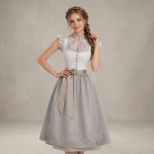 Op de afbeelding: Een traditionele Dirndl-jurk met een wit lijfje met delicate kant en een grijze rok. De jurk heeft een bijpassend grijs schort en een lichtbruine sjerp die in de taille is vastgebonden. De mouwen zijn kort en geplooid.