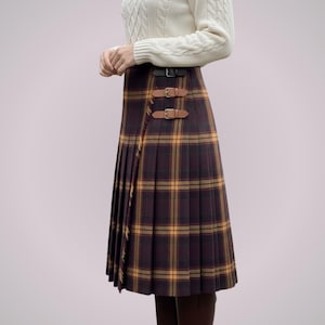 May include: A pleated, knee-length plaid skirt in shades of brown, yellow, and burgundy. The skirt features a wrap-around design with two leather buckle closures. The garment is part of a clothing collection.