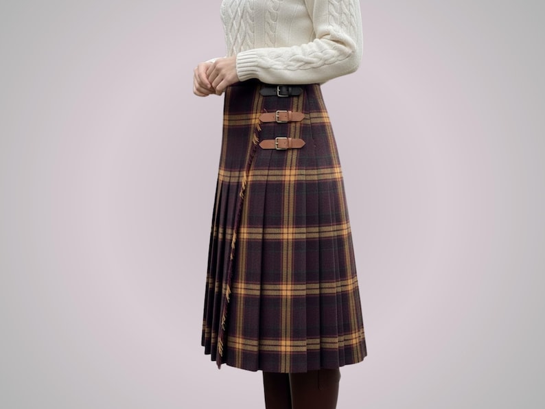 May include: A pleated, knee-length plaid skirt in shades of brown, yellow, and burgundy. The skirt features a wrap-around design with two leather buckle closures. The garment is part of a clothing collection.