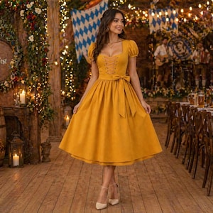 Puede incluir: Un vestido amarillo mostaza con escote cuadrado, mangas cortas y lazo en la cintura. El vestido lo lleva una persona en un ambiente festivo con suelos de madera y luces decorativas.