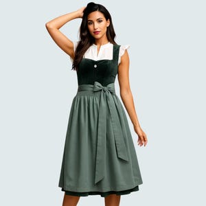 Könnte beinhalten: Ein grünes Dirndlkleid mit einer weißen Bluse. Das Kleid hat ein dunkelgrünes Samtmieder mit einem dekorativen Knopf, eine passende Schürze und einen fließenden Rock. Das Kleid ist knielang.