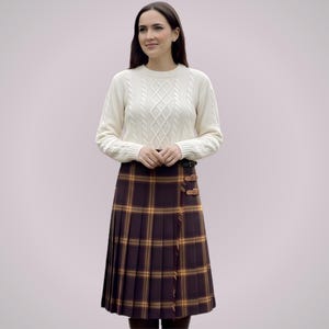 May include: A woman wearing a cream-colored cable-knit sweater and a pleated tartan skirt in shades of brown, yellow, and orange. The skirt features leather buckle details. The outfit is suitable for fall or winter.