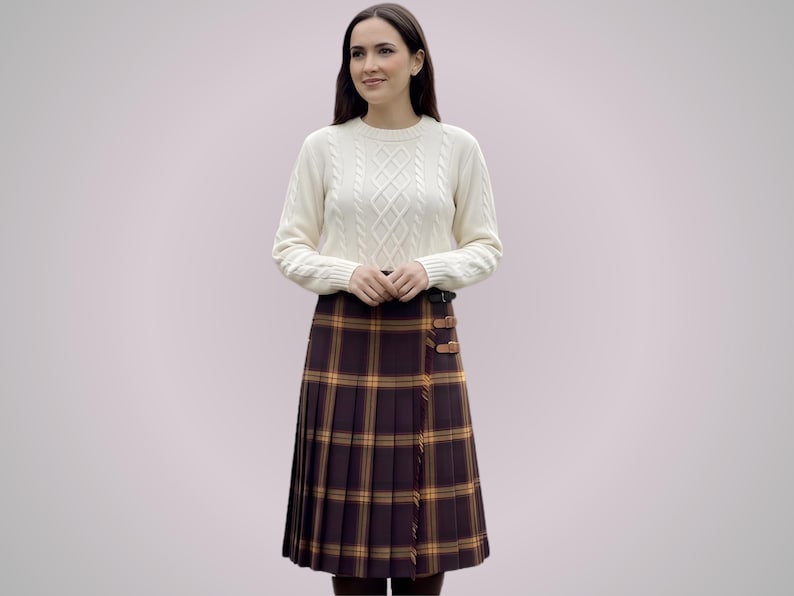 May include: A woman wearing a cream-colored cable-knit sweater and a pleated tartan skirt in shades of brown, yellow, and orange. The skirt features leather buckle details. The outfit is suitable for fall or winter.