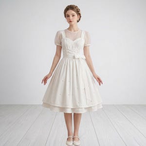 May include: A white knee-length dress with a sheer neckline and short sleeves. The dress features a fitted bodice with a bow detail at the waist and a flared skirt. Small pearl-like embellishments are scattered across the skirt.