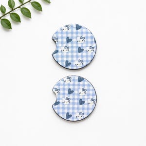 May include: Two round car coasters with a blue and white gingham pattern. Each coaster features a repeating design of blue hearts and a cartoon cat face with a bow. The coasters have a curved cutout for easy removal.