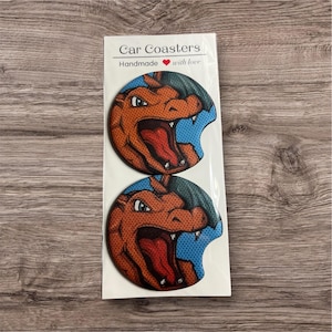 May include: Two car coasters featuring a cartoon dragon design with orange, red, and blue colors. The coasters are round with a curved edge and are packaged together. The packaging reads "Car Coasters Handmade with love."