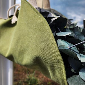 Puede incluir: Una bolsa colgante de tela verde oliva con tejido texturizado, que cuenta con correas de lona color crema y un cierre de cord&oacute;n. La bolsa est&aacute; llena de hojas de eucalipto verde oscuro, mostrando claramente la textura de la tela y las costuras.