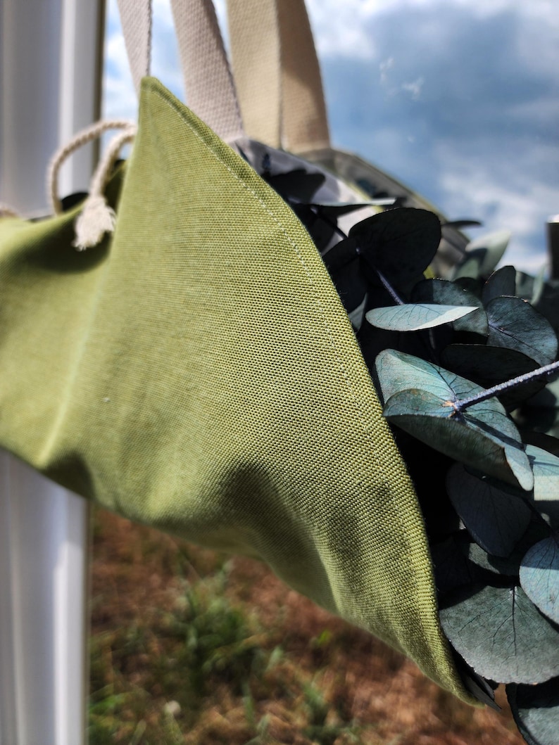 Puede incluir: Una bolsa colgante de tela verde oliva con tejido texturizado, que cuenta con correas de lona color crema y un cierre de cord&oacute;n. La bolsa est&aacute; llena de hojas de eucalipto verde oscuro, mostrando claramente la textura de la tela y las costuras.
