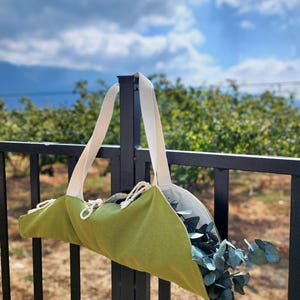 Puede incluir: Un portador de tela verde lima con dos asas de lona color crema y un cierre de cord&oacute;n. El portador est&aacute; lleno de ramas de eucalipto verdes y est&aacute; suspendido de un poste de valla de metal negro contra un cielo azul y follaje verde.