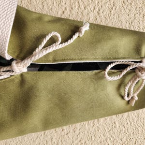 Puede incluir: Una bolsa de tela verde oliva con una abertura central asegurada por dos cordones de cuerda blanca trenzada. El tejido tiene un tramado visible y un acabado liso, con los extremos de los cordones blancos anudados para mantener la bolsa cerrada.