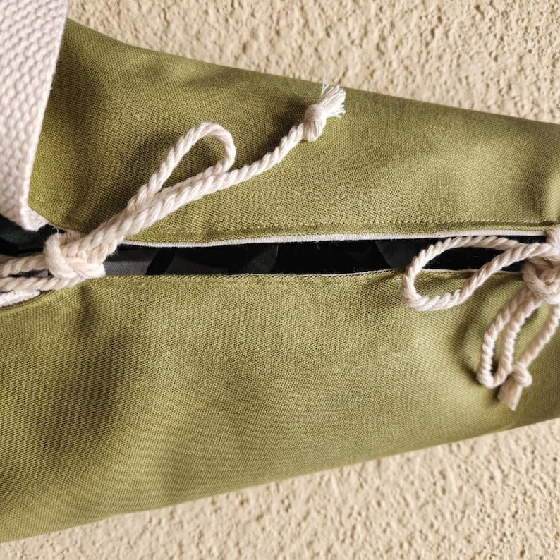 Puede incluir: Una bolsa de tela verde oliva con una abertura central asegurada por dos cordones de cuerda blanca trenzada. El tejido tiene un tramado visible y un acabado liso, con los extremos de los cordones blancos anudados para mantener la bolsa cerrada.