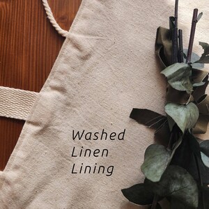 Puede incluir: Un bolso tote beige de tela texturizada con una correa tejida gruesa. El texto "Washed Linen Lining" est&aacute; impreso en negro en la parte frontal. Un manojo decorativo de hojas de eucalipto verdes secas y ramitas de color marr&oacute;n oscuro se sit&uacute;a a la derecha.