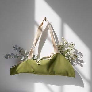Puede incluir: Un bolso tote de lona verde oliva con dos asas de tela color crema y cierre de cord&oacute;n. El bolso est&aacute; relleno con ramas de eucalipto y peque&ntilde;as flores blancas, creando una est&eacute;tica natural con una textura de tela claramente visible.