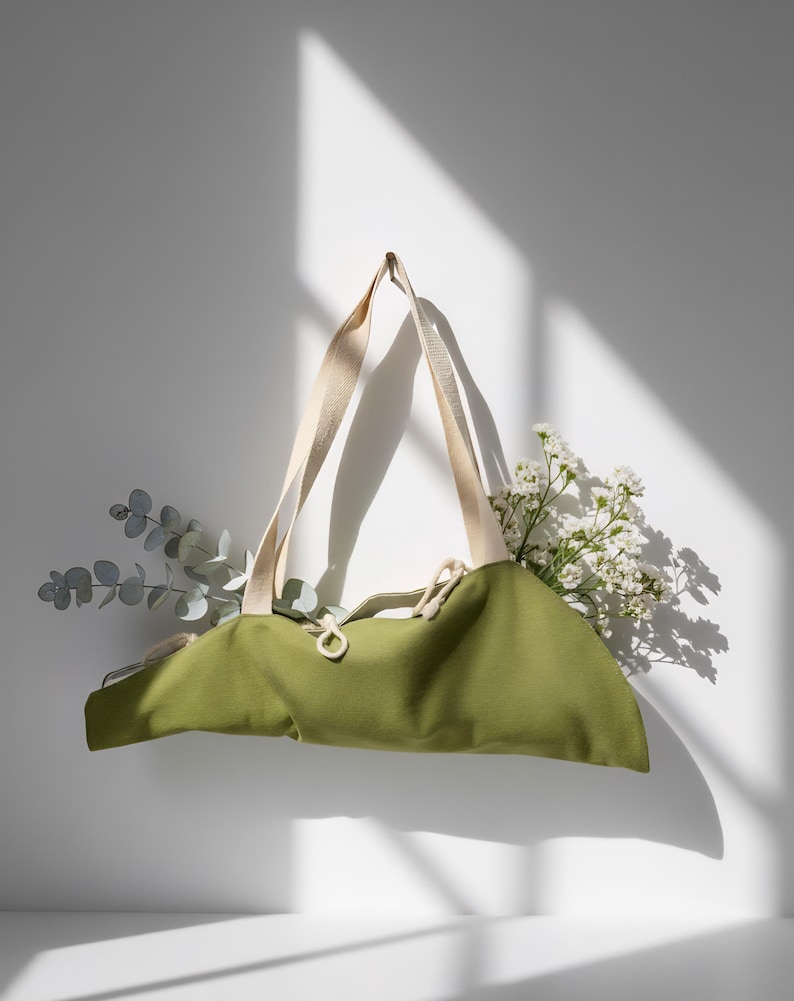 Puede incluir: Un bolso tote de lona verde oliva con dos asas de tela color crema y cierre de cord&oacute;n. El bolso est&aacute; relleno con ramas de eucalipto y peque&ntilde;as flores blancas, creando una est&eacute;tica natural con una textura de tela claramente visible.