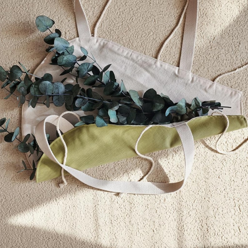 Puede incluir: Dos bolsas de tela con asas planas y cordones, una en color beige claro y otra en verde oliva apagado, superpuestas con una larga rama de eucalipto con hojas redondeadas de color verde oscuro que descansa sobre el centro de las bolsas.
